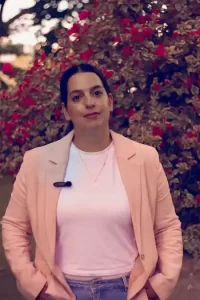 אירית שלום, מאמנת נשים לאחר אובדן, עומדת בחוץ מול שיח פרחים ורודים לובשת ג'קט אפרסק