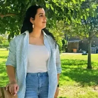 אירית שלום עומדת בחוץ מתחת לעץ בגינה, לובשת חולצה לבנה וקרדיגן בהיר